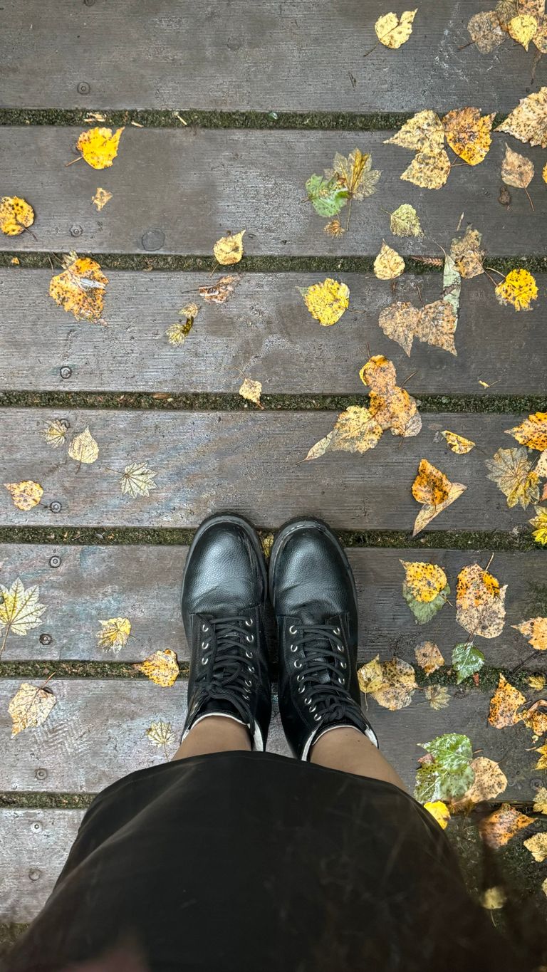 Top-down view of black boots on wooden planks with scattered autumn leaves.